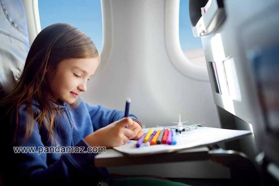 عکس یک دختربچه در هواپیما در حال پرواز در آسمان. در تصویر دختر کوچولو با بلوز آبی رنگ بر روی صندلی هواپیما با آرامش و با لبخند نشسته و در حال نقاشی روی کاغذ با ماژیک های رنگی است