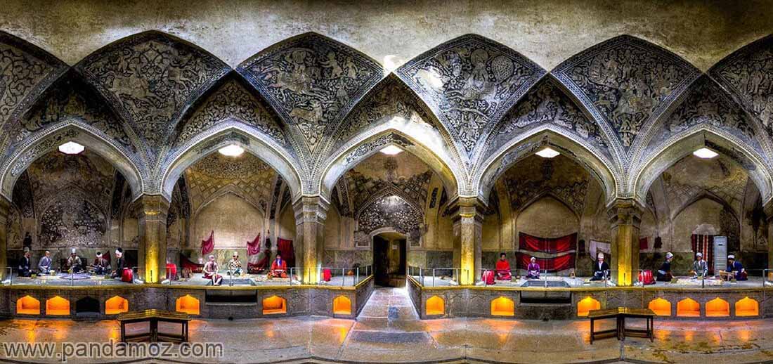 عکس حمام وکیل شیراز با طاق ها و گچ بری های بسیار زیبا در سقف و نمای کامل حمام با چراغ های و نورها. در تصویر مجسمه های مومی دلاکها، مشتریان و مردم در نیم طبقه ها و تاقچه های حمام دیده می شوند