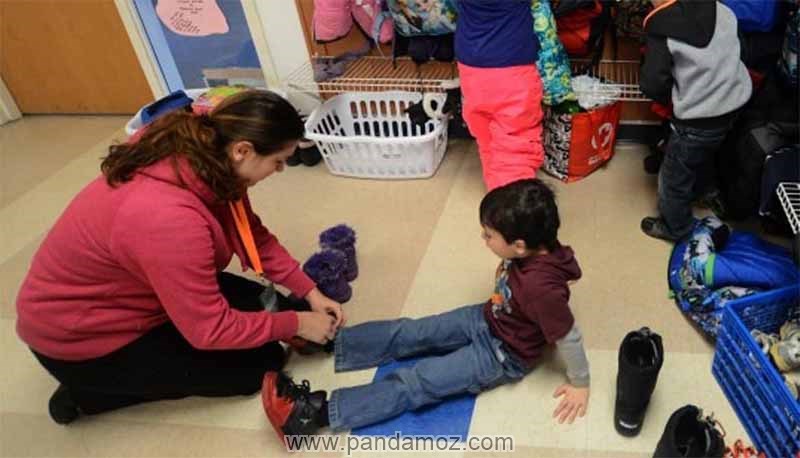 عکس مهد کودک و مربی زن مهد کودک که روی زمین نشسته و در حال بستن بندهای پوتین ها و کفش پسر بچه است. در تصویر دو تن از بچه ها و وسائل آنها دیده می شود
