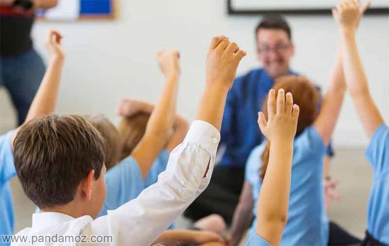 عکس کلاس درس و معلم آقا. تصویر روی دستهای دانش آموزان از پشت سر که برای پاسخ دادن به سوال معلم بلند شده، زوم شده و معلم مرد به صورت مات رو به تصویر دیده می شود و پیراهن آبی بر تن دارد