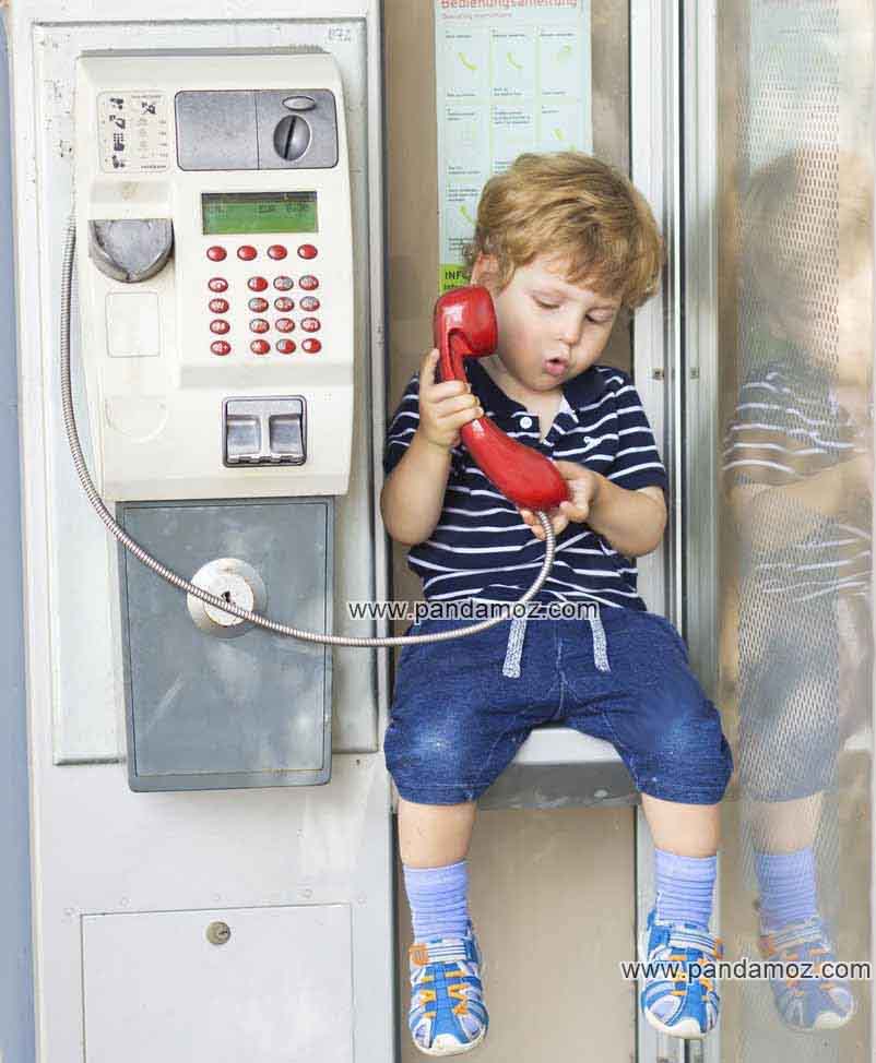 عکس پسر بچه در تلفن همگانی که بر روی سکوی داخلی کیوسک تلفن عمومی نشسته و گوشی تلفن قرمز رنگ در دست دارد