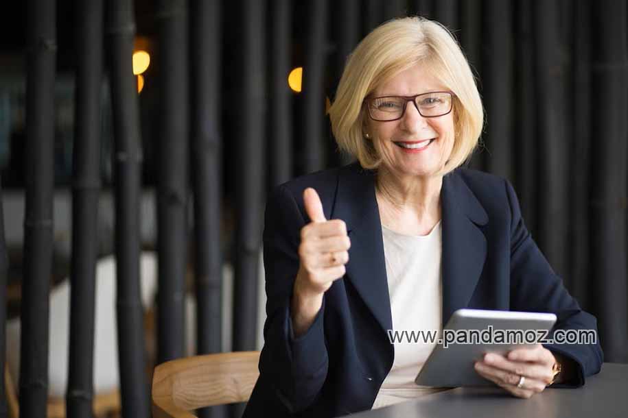 عکس یک پیرزن با عینک در پشت میز با تبلت در دست. در تصویر پیرزن با موهای بلوند و کت سرمه ایی در جایی مثل کافی شاپ با لبخندی بر لب نشسته و  دست راست خود را به علامت لایک بالا برده است