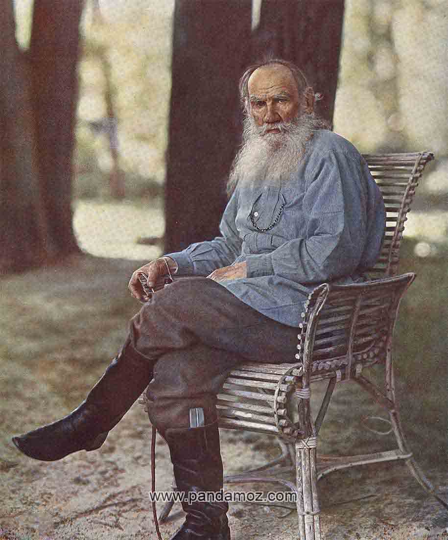 عکس رنگی شده لئو تولستوی نویسنده روسی در پارک که روی نیمکت پارک نشسته و پشت سرش درختان پارک و زمین چمن دیده می شود. در تصویر تولستوی ریش سفید بلندی دارد و چکمه پوشیده است