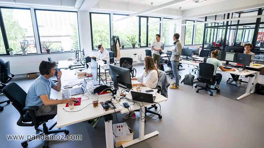 عکس نمای داخلی یک شرکت و کارمند هایی که مشغول کار هستند شامل میزهای کار و کامپیوترها و افرادی که در حال صحبت و همفکری باهم هستند