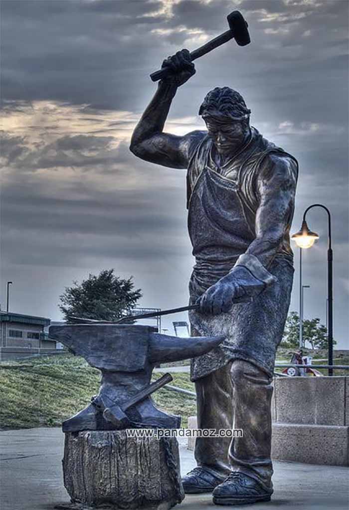 عکس مجسمه آهنگر که روپوش چرمی آهنگری بر تن دارد و پتکی را با دست بلند کرده و در حال فرود آوردن بر روی آهن گداخته روی سندان آهنگری است. در پشت تصویر فضای پارک و محل تندیس آهنگر دیده می شود