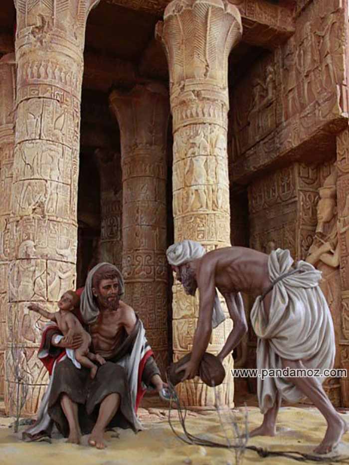 عکس مجسمه مردی رنگین پوست که بسته گرانبها و گران قیمتی را در حال خمیده و در دست به مردی دیگری که بر زمین نشسته می دهد. در تصویر مرد نشسته بچه ایی در بغل دارد و زیر کاخی با ستون های بلند و افراشته نشسته. روی سنگهای دیوار و ستون کاخ تصاویری حکاکی شده