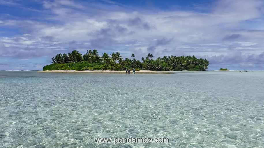 عکس یک جزیره زیبا در وسط اقیانوس بادرختان بلند و سرسبز و احاطه آب کامل دور جزیره، چند نفر در ساحل جزیره از دور دیده می شوند. ابرها در آسمان آبی و موج های کوتاه دریا جلوه خاصی دارند