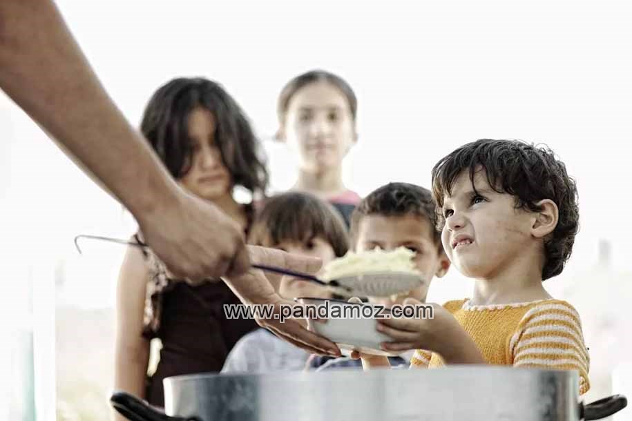 عکس غذا دادن به تعدادی بچه نیازمند. در تصویر  قابلمه و دستی که با کف گیر در ظرف یک پسربچه غذا می ریزد و پشت سر او تعدادی بچه دختر و پسر در صف غذا دیده می شوند