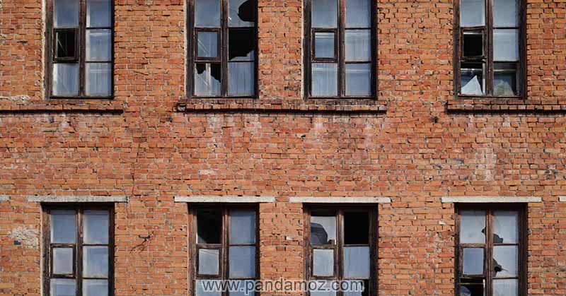 عکس نمای ساختمان چند طبقه آچری قدیمی که شیشه های پنجره های آن شکسته شده در تصویر آجرهای قهوه ایی نمای ساختمان نیز نامنظم و پنجره ها چوبی است
