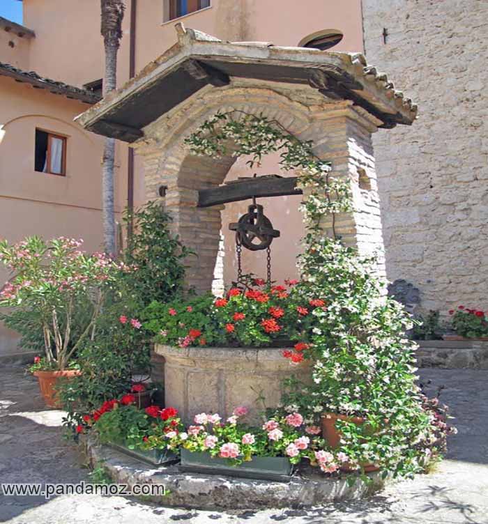 عکس یک چاه قدیمی با سایه بان آجری که با گلها و گیاهان اطراف پوشیده شده. در تصویر قرقره و زنجیر چاه و پایه آجری آن دیده می شود. گل های شمعدانی در اطراف چاه دیده می شوند. پشت چاه ساختمان مسکونی قرار دارد