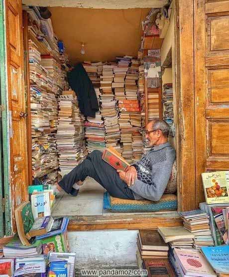 عکس کتاب فروش کتابهای دست دوم که فارغ از دنیا در حال مطالعه کتاب است. در تصویر او در چهارچوب در نشسته و پاهایش را کمی خم کرده