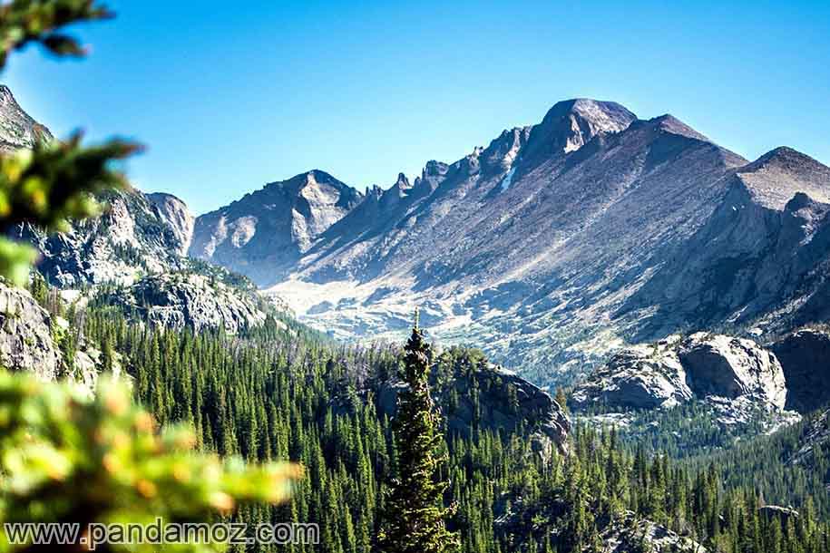 عکس پوستر کوه زیبا و دره بین کوهها و گیاهان و درختان سرسبز در دامنه کوه. تصویر منظره کوهستانی زیبا را نشان می دهد که در برخی از نواحی کوه همچنان برف وجود دارد