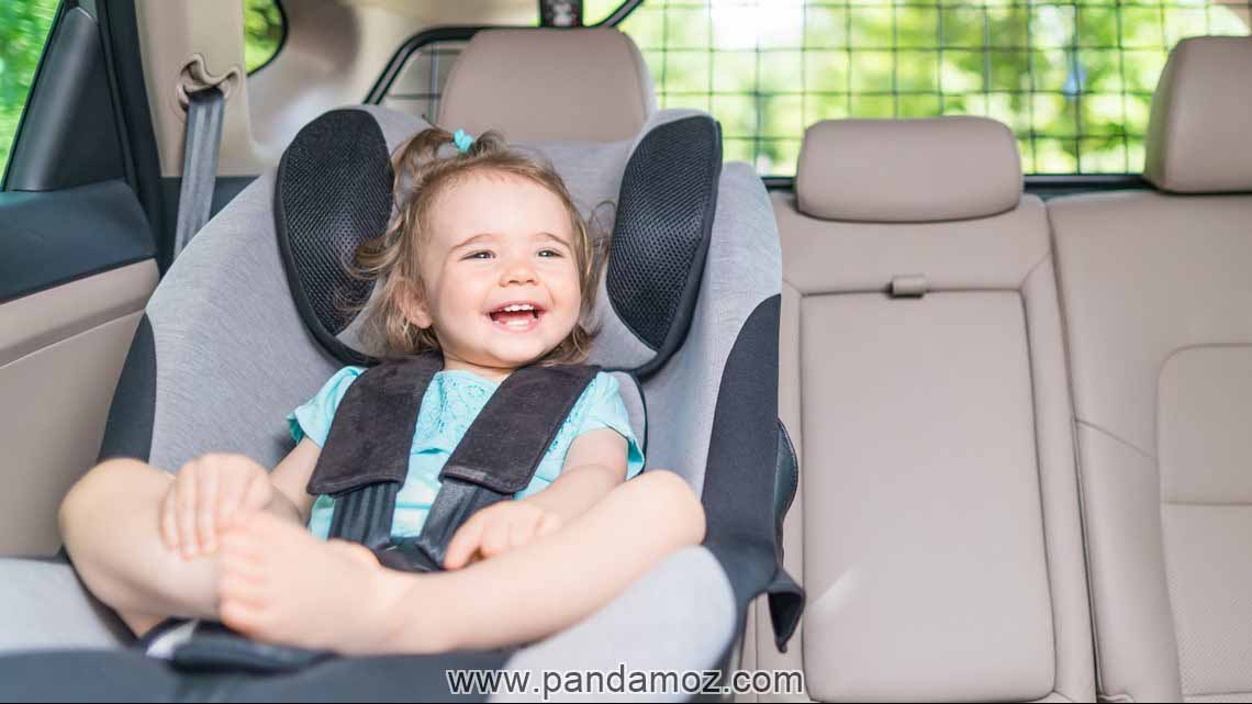 عکس کودک نشسته روی صندلی جلو ماشین که با کمربند و نگهدارنده کودک بسته شده. در تصویر کودک خندان و شاد است