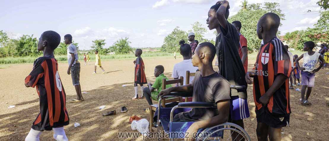 عکس تعداد زیادی پسربچه سیاه پوست آفریقایی  که به بازی فوتبال در زمین خاکی نگاه می کنند. در تصویر یکی از پسرها معلول بوده و روی ویلچر نشسته است.