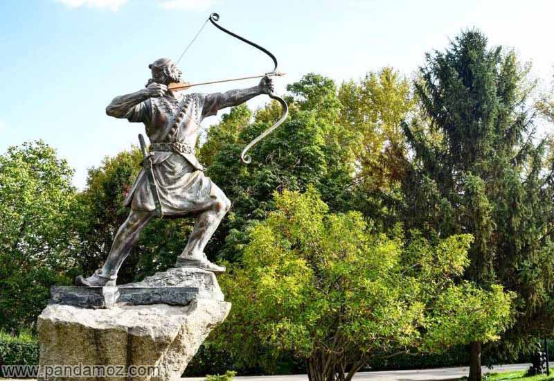 عکس مجسمه برنزی شکارچی به صورت ایستاده. در تصویر تندیس مرد کمانگیر، تیری را در چله کمان گذاشته و کشیده و آماده برای رها کردن و پرتاب تیر است. مجسمه در پارکی قرار داشته و درختان سرسبز اطراف آن را احاطه کرده اند