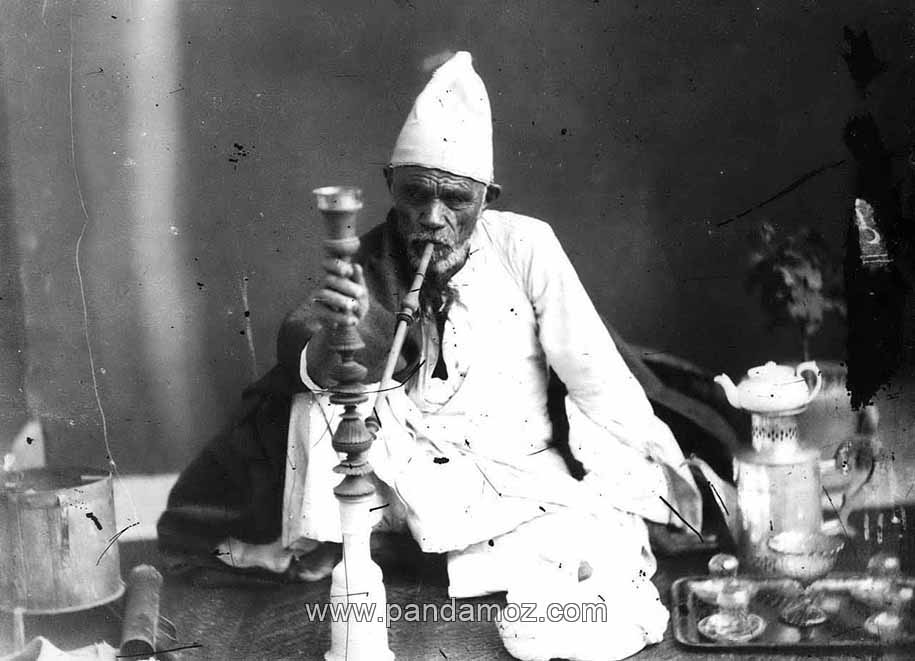 عکس سیاه و سفید از کریم شیره ای دلقک مشهور دربار ناصرالدین شاه قاجار در حال کشیدن قلیان. در تصویر او کلاهی نمدی و قدیمی بر سر دارد و سماور با قوری چینی روی آن در کنارش دیده می شود