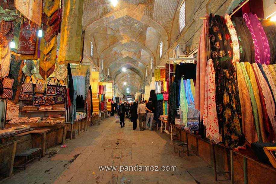 عکس زیبا و رنگارنگ از داخل بازار وکیل شیراز با طاق های بلند بازار و حجره های فروشندگان. در تصویر مغازه هایی که لباس های رنگی محلی عشایر را می فروشند و همچنین صنایع دستی استان فارس دیده می شود
