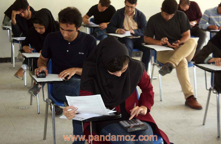 عکس یک کلاس دانشگاه که دانشجویان در حال امتحان دادن هستند. در تصویر دختر دانشجو در جلو و پسرهای دانشجو در پشت بر روی صندلی های دسته دار در حال پاسخ دادن به ورقه امتحانی دیده می شوند. بعضی ها ماشین حساب در دست دارند