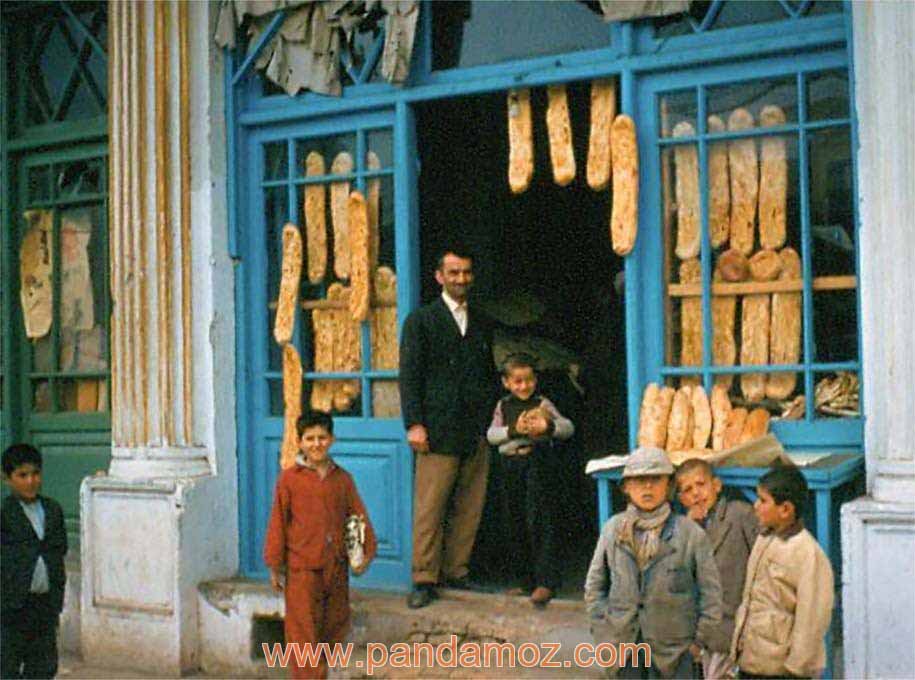 عکس مغازه نانوایی در تهران قدیم. تصویر رنگی شده از یک عکس سیاه و سفید قدیمی از یک مغازه بربری فروشی در تهران. در جلوی در ورودی مغازه یک پسربچه و مردی با لباس مرتب ایستاده و چند پسر بچه نیز بیرون از مغازه دیده می شوند. درب مغازه چوبی بوده و به رنگ آبی است 